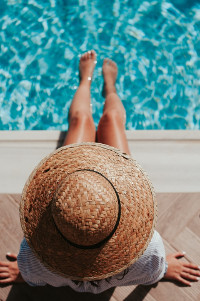 Frau mit Sonnenhut relaxt am eigenen Swimming-Pool mit den Füßen im Wasser